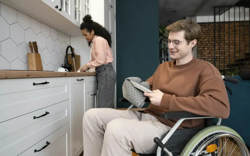 A smiling young man in a wheelchair reads a tablet in a kitchen while a woman prepares food at the bench behind him
